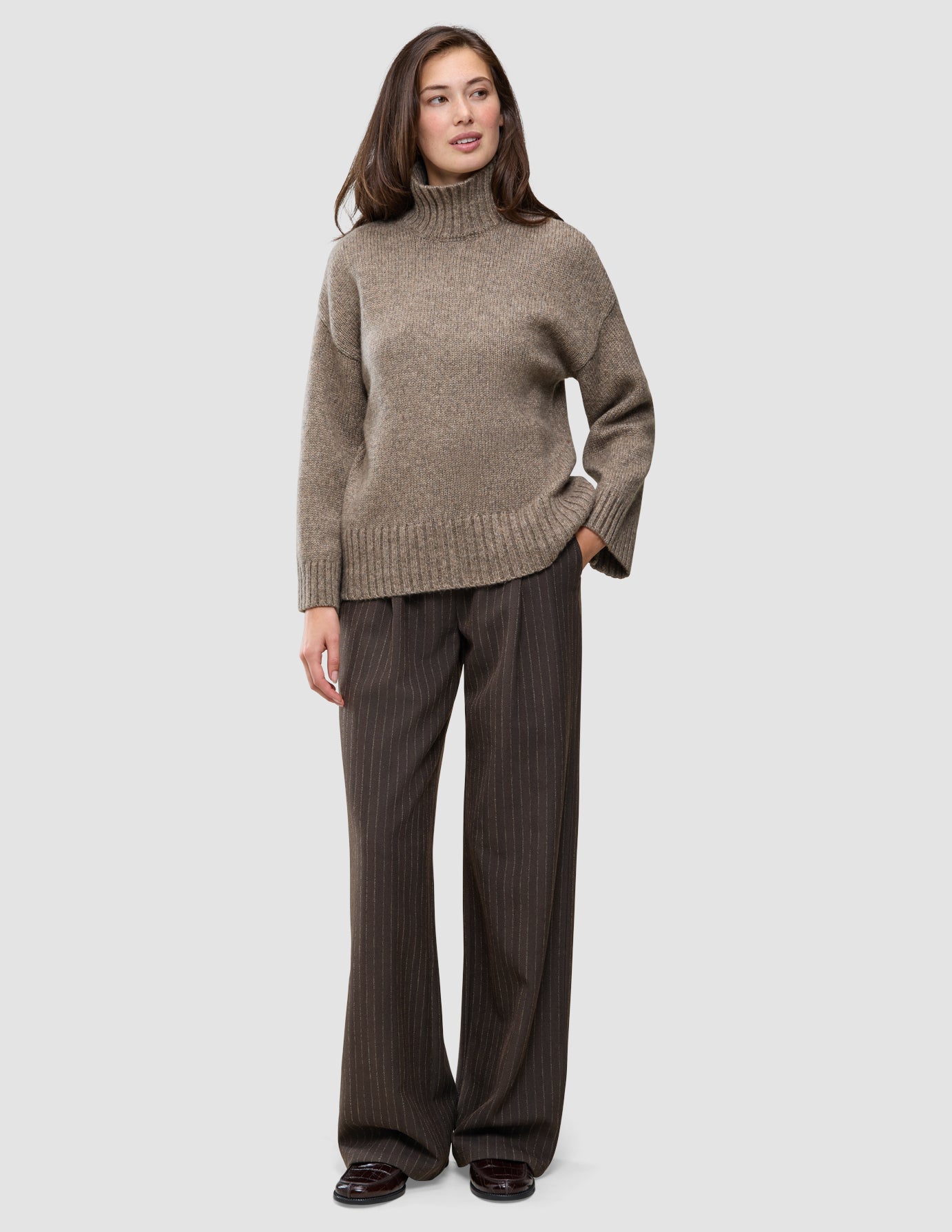 Eine Frau trägt einen dicken, weichen Wollpullover mit Rollkragen in Taupe-Melange und eine braune Nadelstreifenhose.