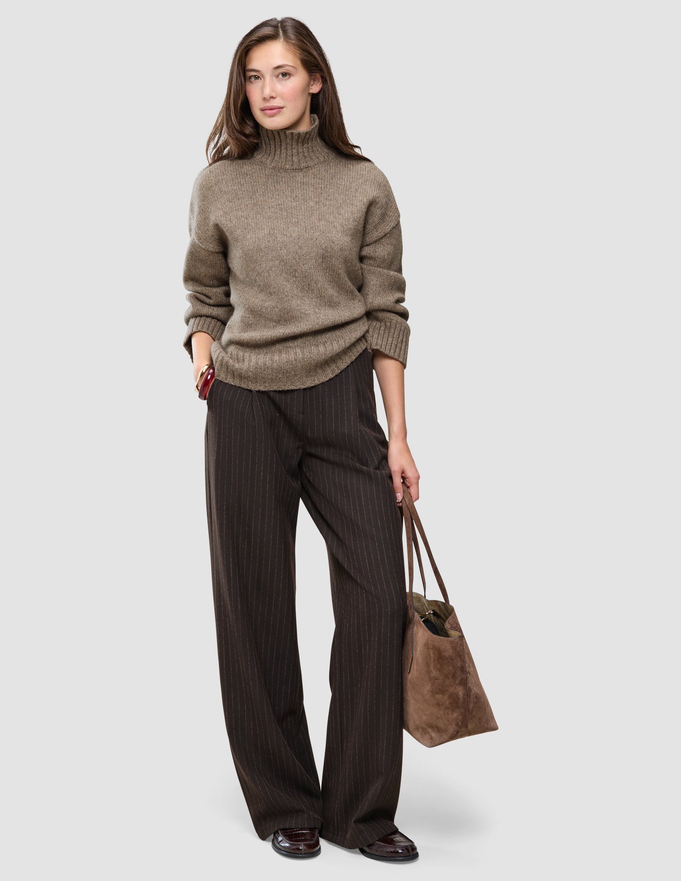 Eine Frau trägt einen taupefarbenen, melierten Damen-Rollkragenpullover aus dicker, weicher Wolle und eine dunkelbraune Nadelstreifenhose sowie eine braune Tragetasche.