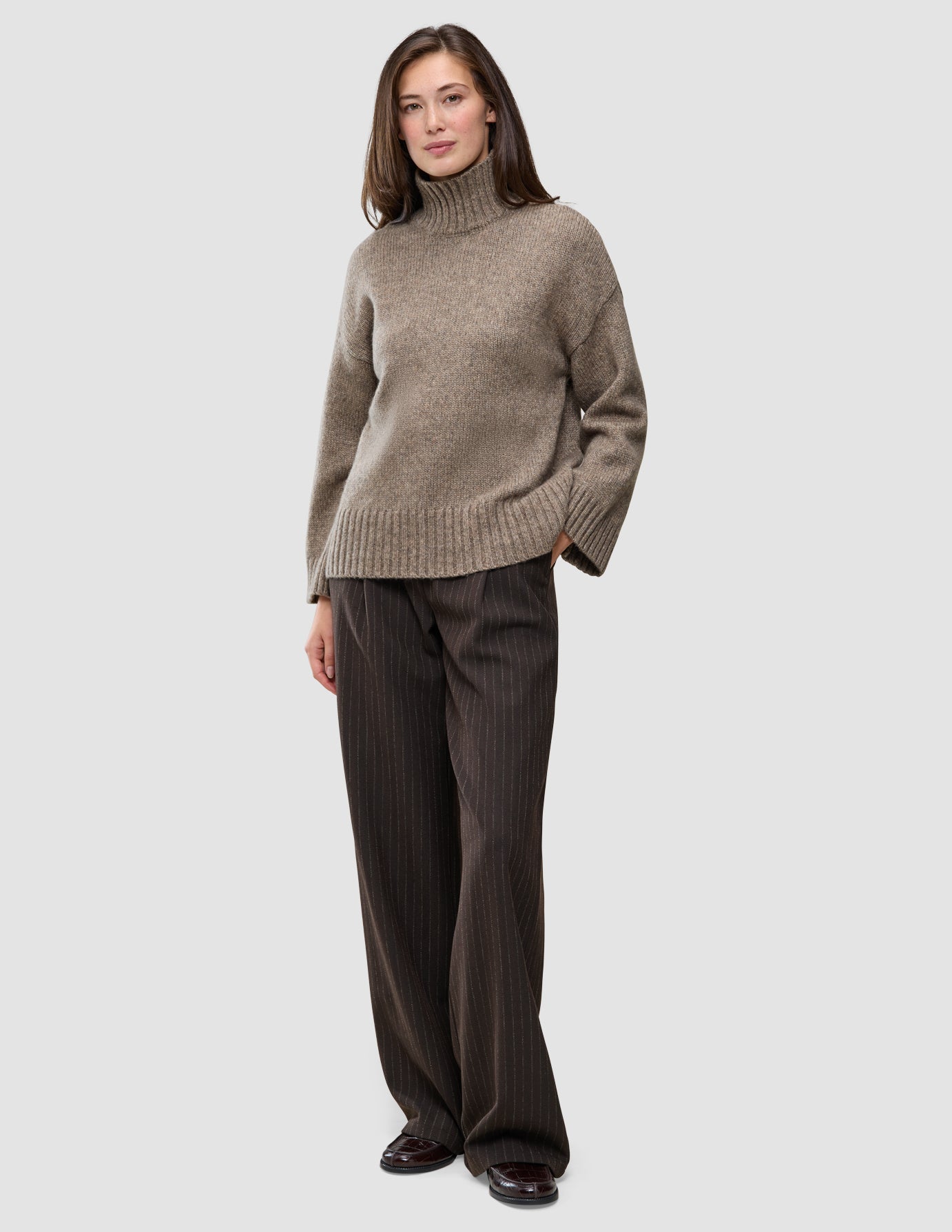 Eine Frau, die einen taupefarbenen, melierten, grob gestrickten, weichen Woll-Rollkragenpullover für Damen und eine weit geschnittene Nadelstreifenhose trägt.