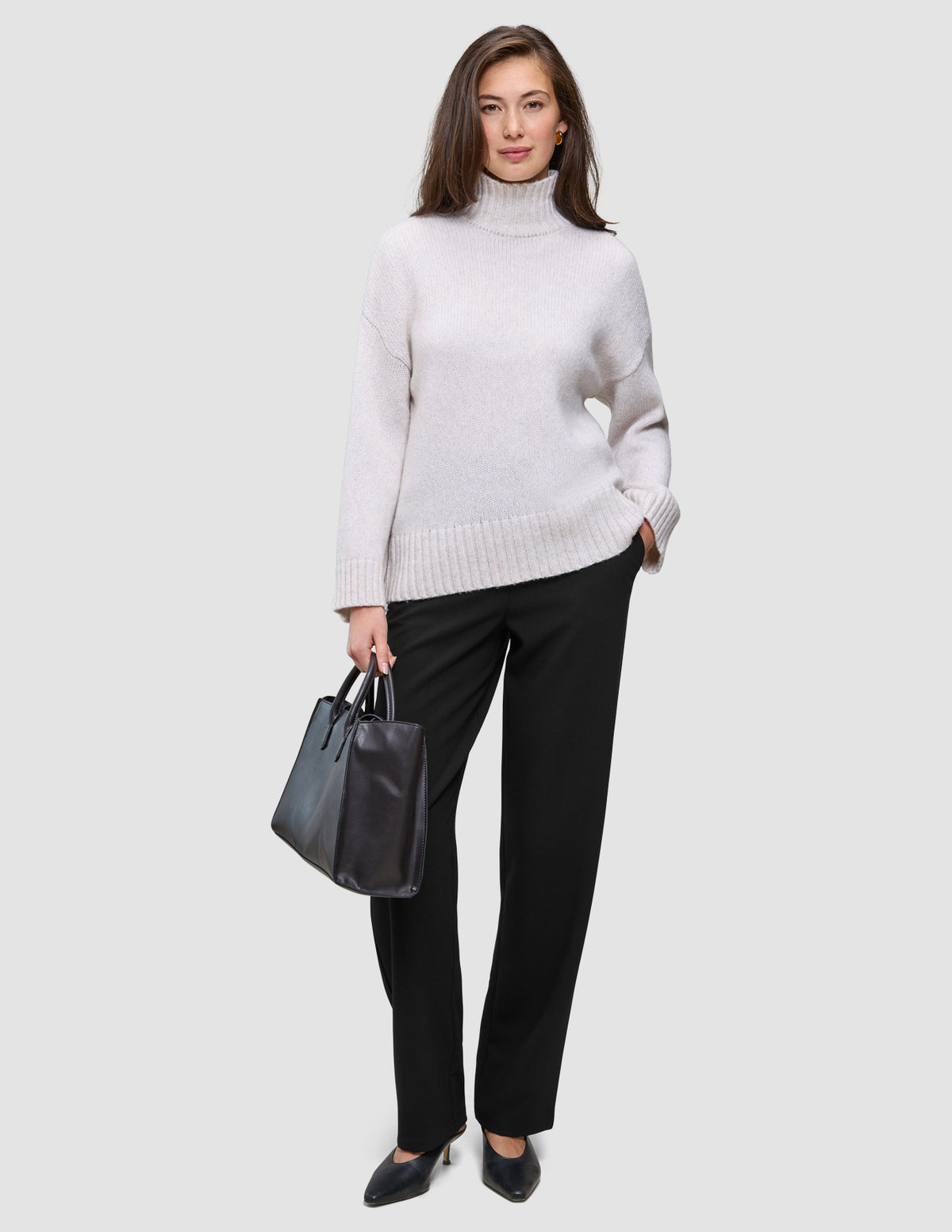 Eine Frau, die einen beigen Rollkragenpullover und eine schwarze Women GO2 Pants Straight trägt und eine schwarze Handtasche in der Hand hält.
