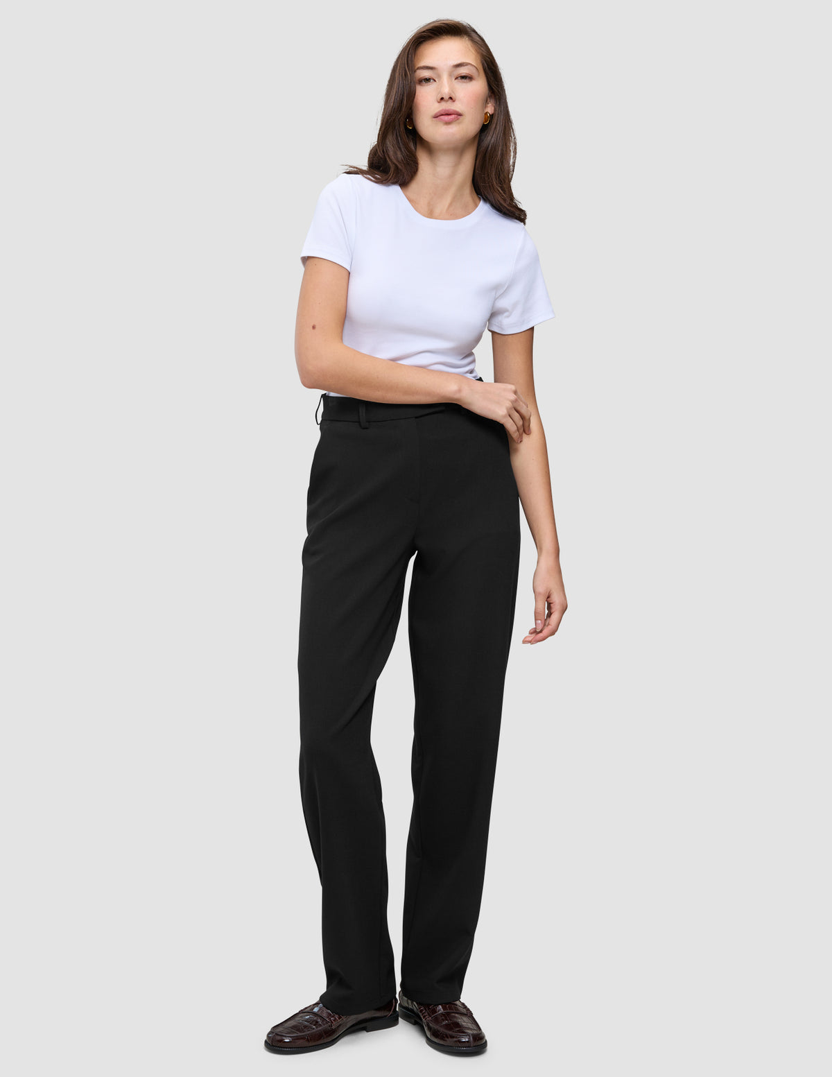 Eine Frau in einem weißen T-Shirt mit Rundhalsausschnitt, einer schwarzen Women GO2-Hose in gerader Passform und dunkelbraunen Loafern, die ein minimalistisches skandinavisches Design widerspiegeln.
