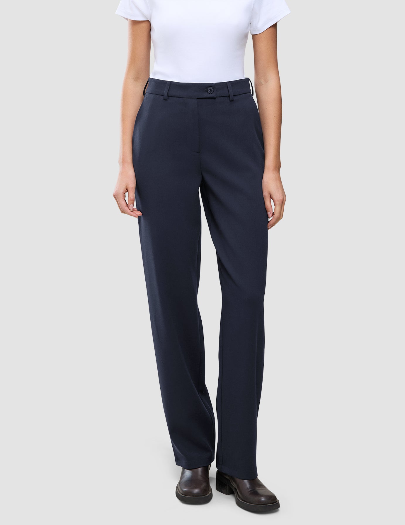 Eine Person trägt ein weißes T-Shirt, das in die Women GO2 Pants Straight Blue Nights mit Knopfverschluss gesteckt ist, dazu dunkelbraune Schuhe im minimalistischen skandinavischen Design.