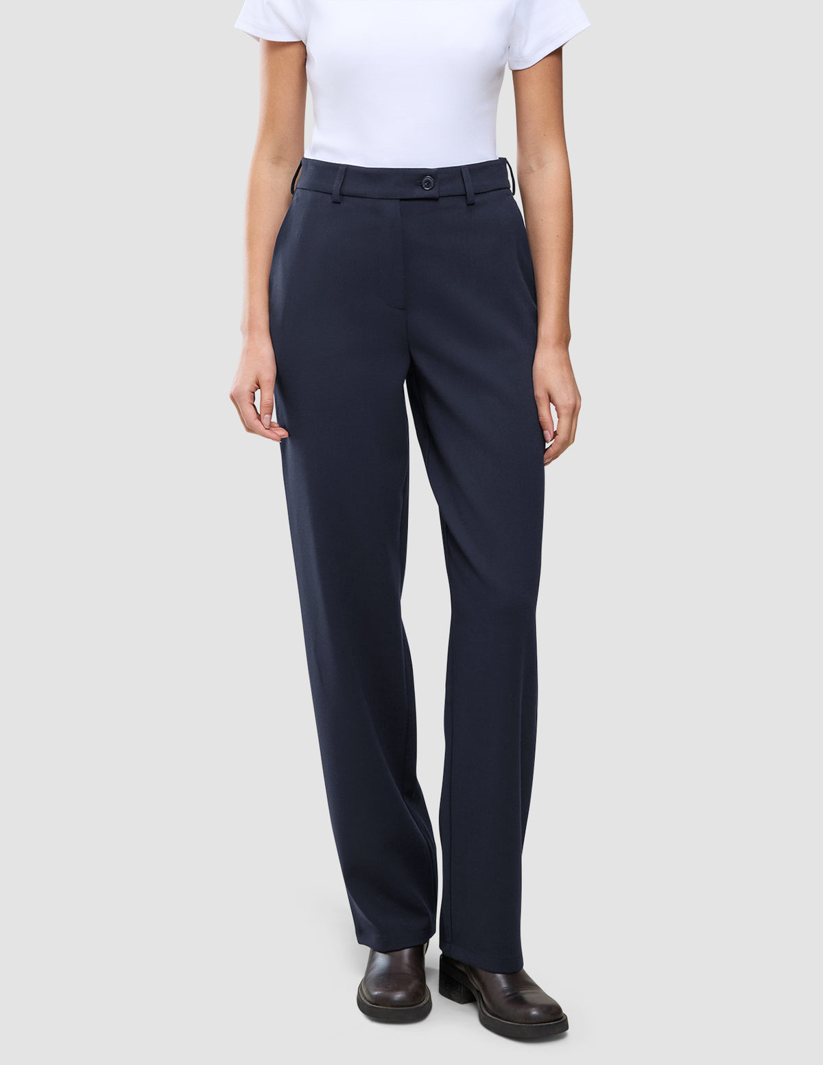 Eine Person trägt ein weißes T-Shirt, das in die Women GO2 Pants Straight Blue Nights mit Knopfverschluss gesteckt ist, dazu dunkelbraune Schuhe im minimalistischen skandinavischen Design.