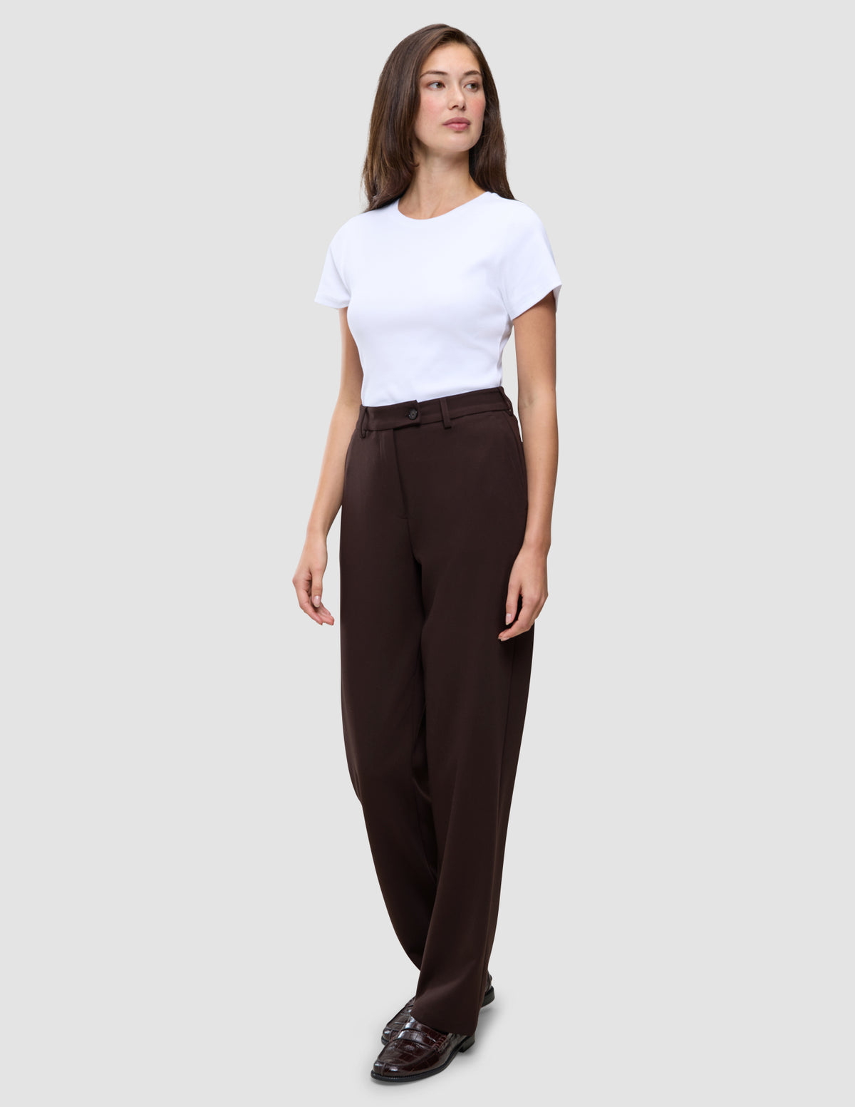 Eine Frau, die ein weißes T-Shirt, eine Women GO2 Pants Straight Mocha und dunkle Loafer trägt, die ein klares, skandinavisches Design widerspiegeln.