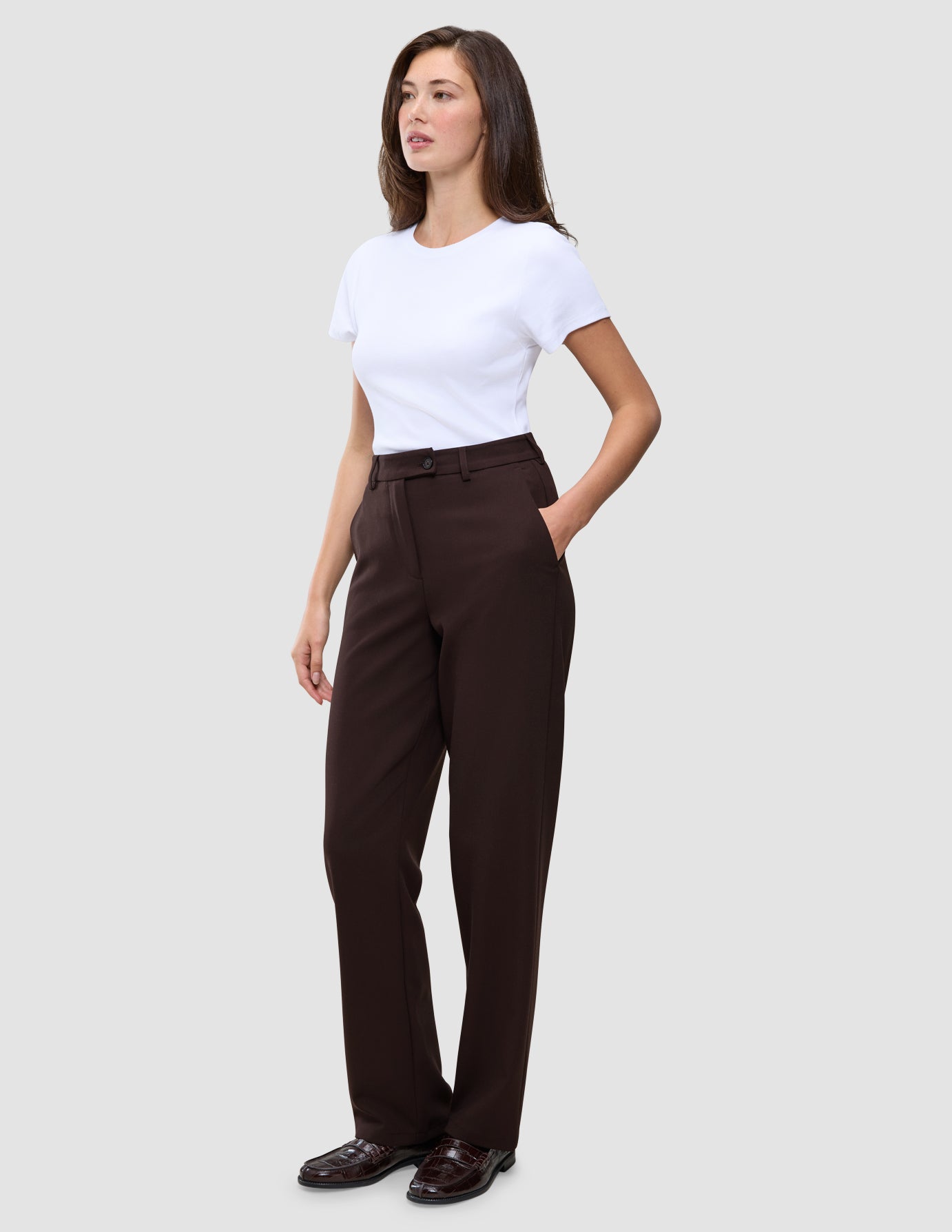 Eine Person, die das minimalistische skandinavische Design verkörpert: Sie trägt ein weißes T-Shirt, die Women GO2 Pants Straight Mocha in Dunkelbraun mit Knopfverschluss und dunkle Loafer.