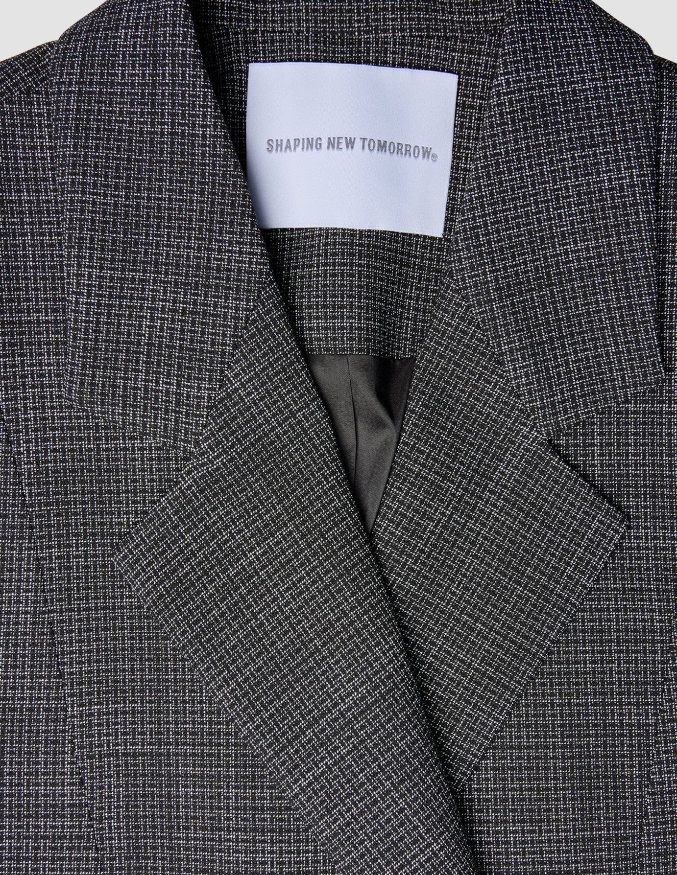 Nahaufnahme eines Women Nara Blazer Fitted Black Check mit einem maßgeschneiderten schwarz-weißen Karomuster und einem weißen Etikett im Nackenbereich, auf dem SHAPING NEW TOMORROW steht.