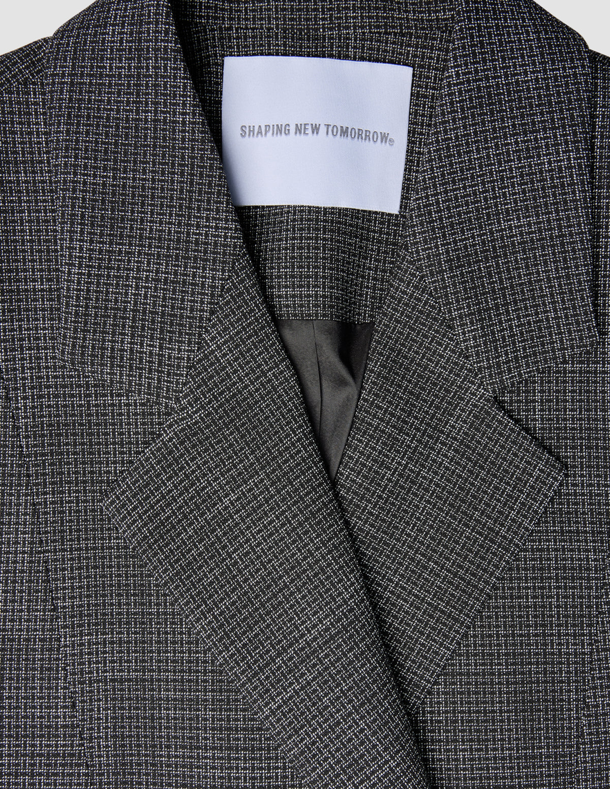 Nahaufnahme eines Women Nara Blazer Fitted Black Check mit einem maßgeschneiderten schwarz-weißen Karomuster und einem weißen Etikett im Nackenbereich, auf dem SHAPING NEW TOMORROW steht.