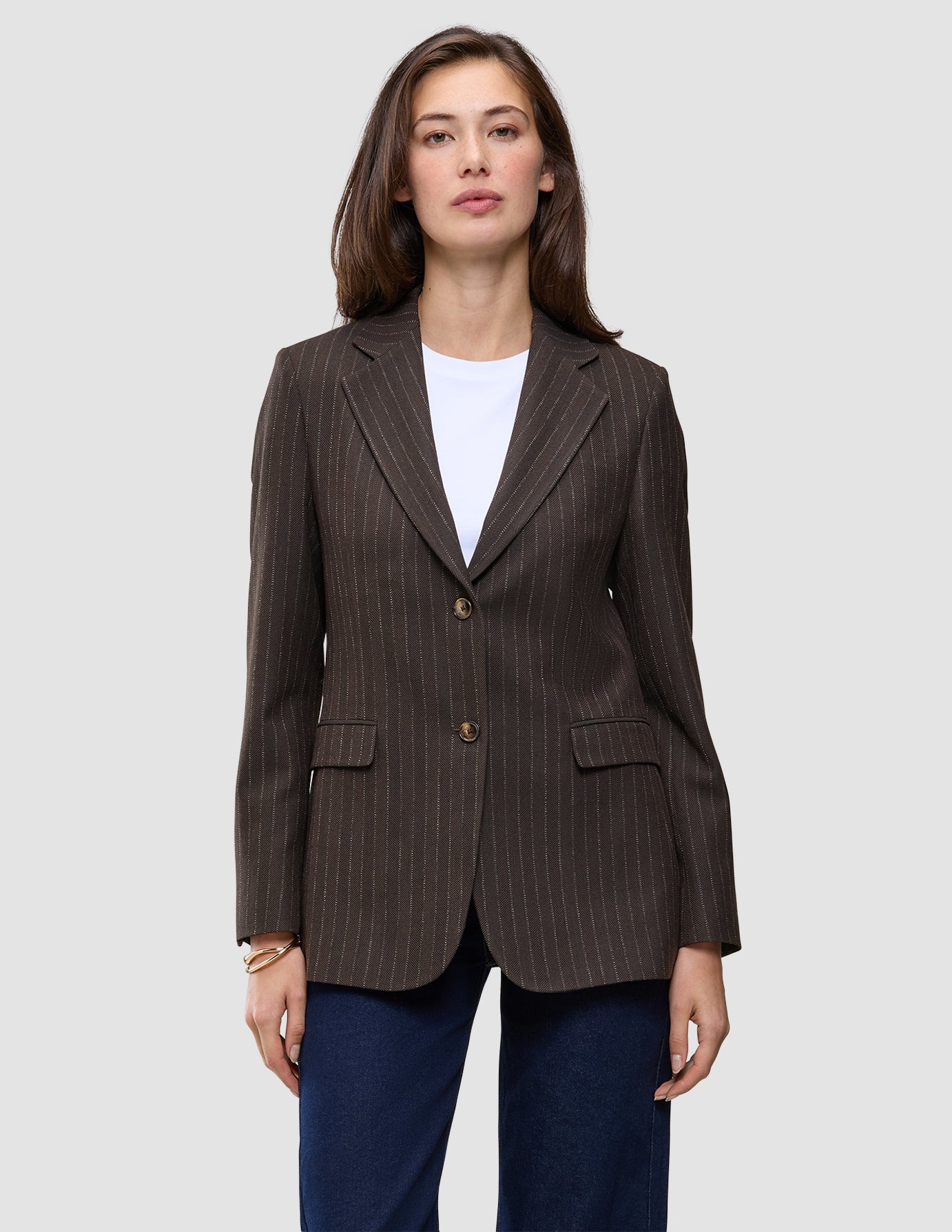 Eine Frau trägt einen Women Serene Blazer Regular Walnut Herringbone, einen braunen, zweifarbigen Blazer mit Nadelstreifen und zwei Knöpfen, über einem weißen Hemd und dunkelblauen Jeans.