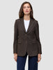 Eine Frau trägt einen Women Serene Blazer Regular Walnut Herringbone, einen braunen, zweifarbigen Blazer mit Nadelstreifen und zwei Knöpfen, über einem weißen Hemd und dunkelblauen Jeans.