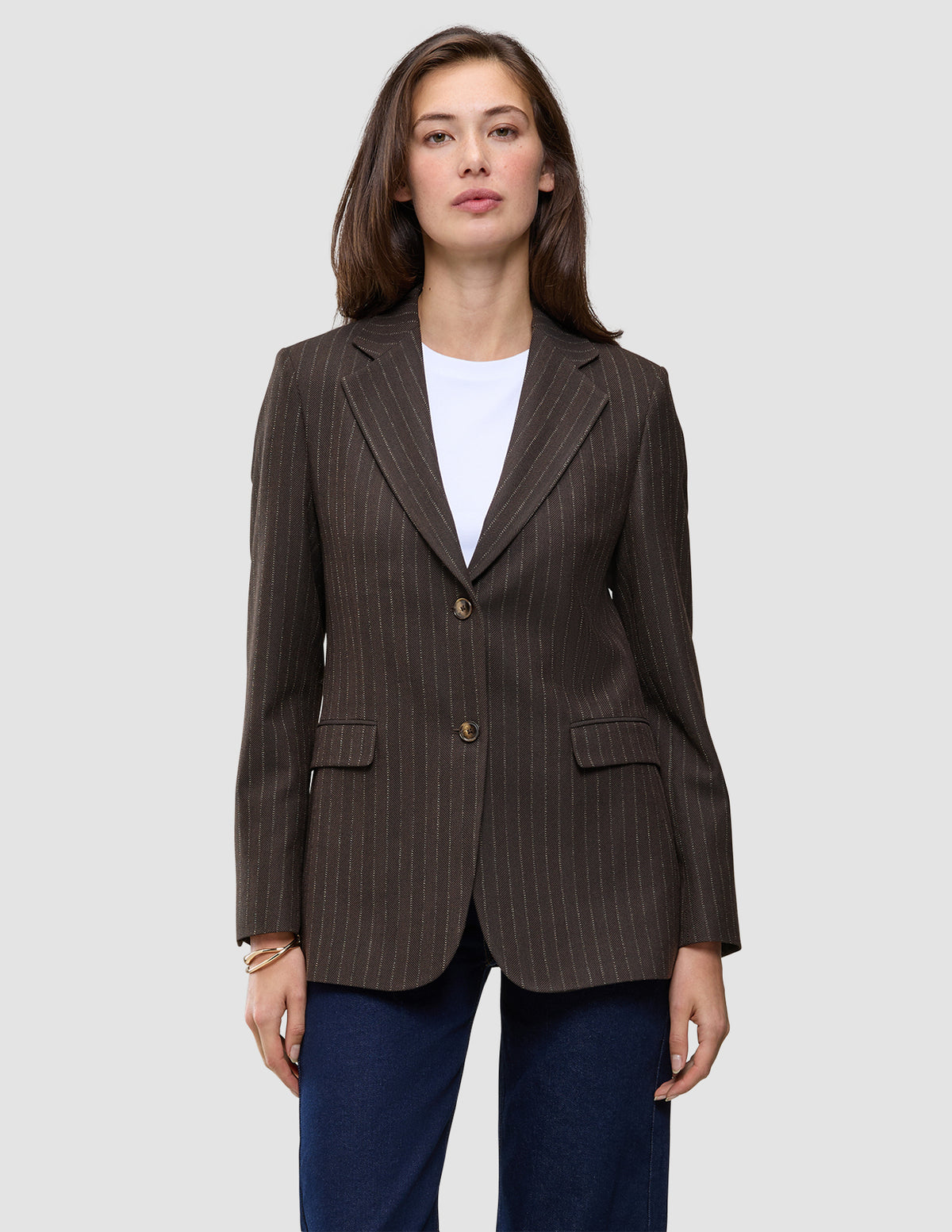 Eine Frau trägt einen Women Serene Blazer Regular Walnut Herringbone, einen braunen, zweifarbigen Blazer mit Nadelstreifen und zwei Knöpfen, über einem weißen Hemd und dunkelblauen Jeans.