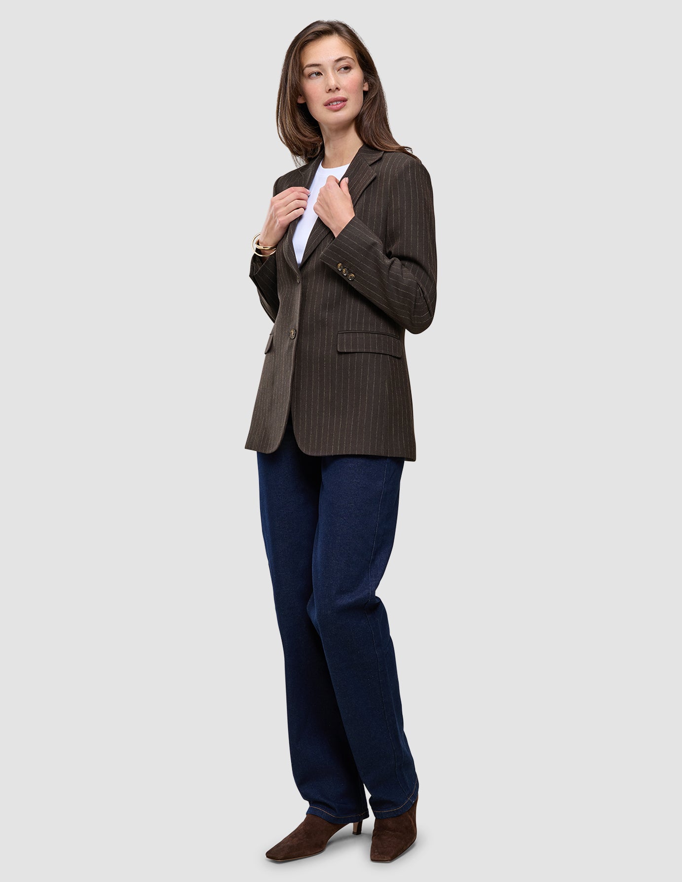 Eine Person trägt den Women Serene Blazer Regular Walnut Herringbone über einem weißen T-Shirt, dunkelblauen Jeans und braunen Stiefeletten.