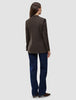 Eine Person, von hinten gesehen, die einen Women Serene Blazer Regular Walnut Herringbone, eine dunkelblaue Jeans und braune Schuhe trägt.