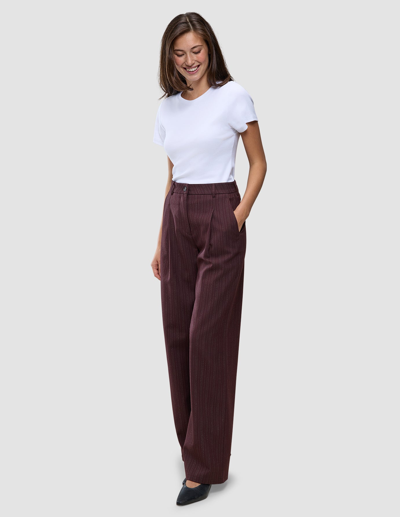 Eine Frau trägt ein weißes T-Shirt und die Women Serene Pants Wide Berry Pinstripe – eine hoch taillierte, weit geschnittene Hose in Beerenfarbe mit Nadelstreifen im skandinavischen Design.