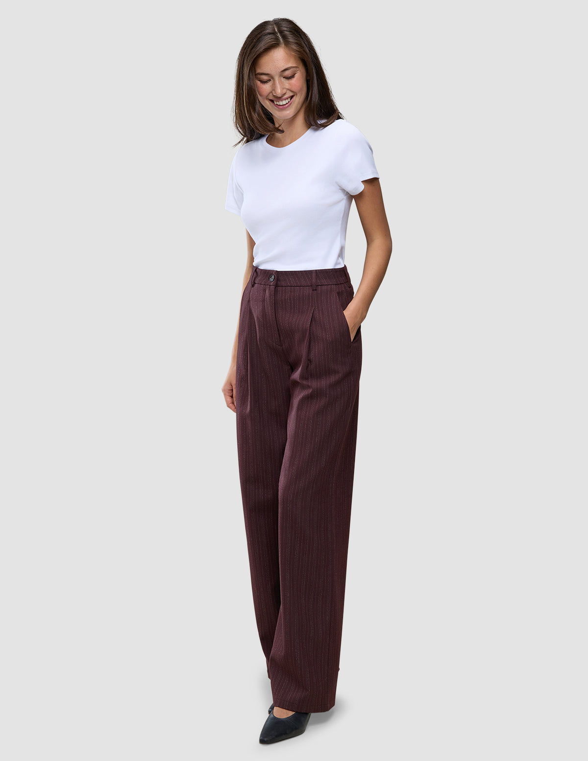 Eine Frau trägt ein weißes T-Shirt und die Women Serene Pants Wide Berry Pinstripe – eine hoch taillierte, weit geschnittene Hose in Beerenfarbe mit Nadelstreifen im skandinavischen Design.