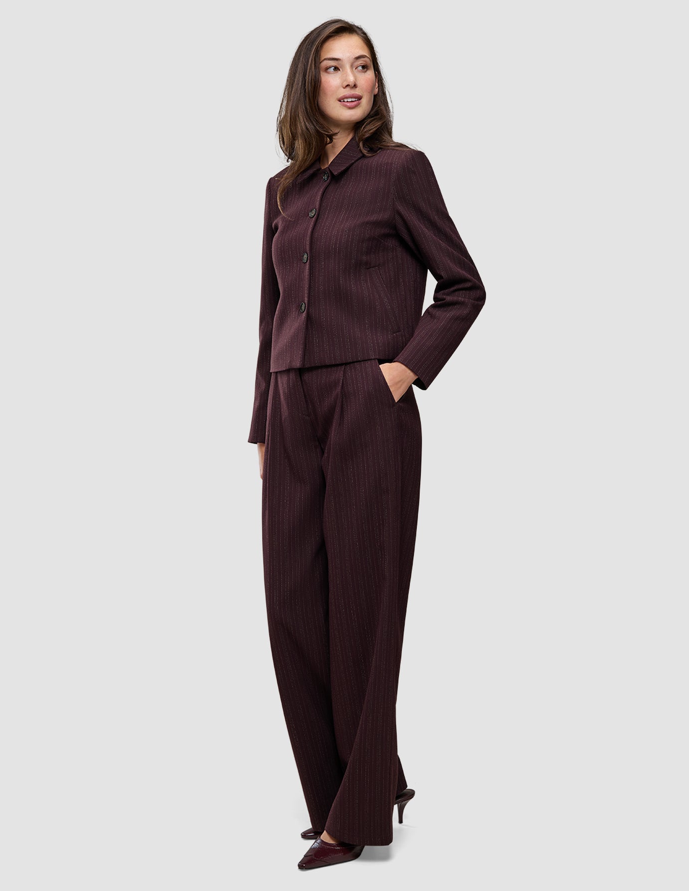 Eine Frau trägt die Women Serene Short Jacket Berry Pinstripe, eine kastenförmige, dunkelviolette Jacke mit Nadelstreifen, kombiniert mit einer passenden Hose mit hoher Taille im schlichten, skandinavischen Design.