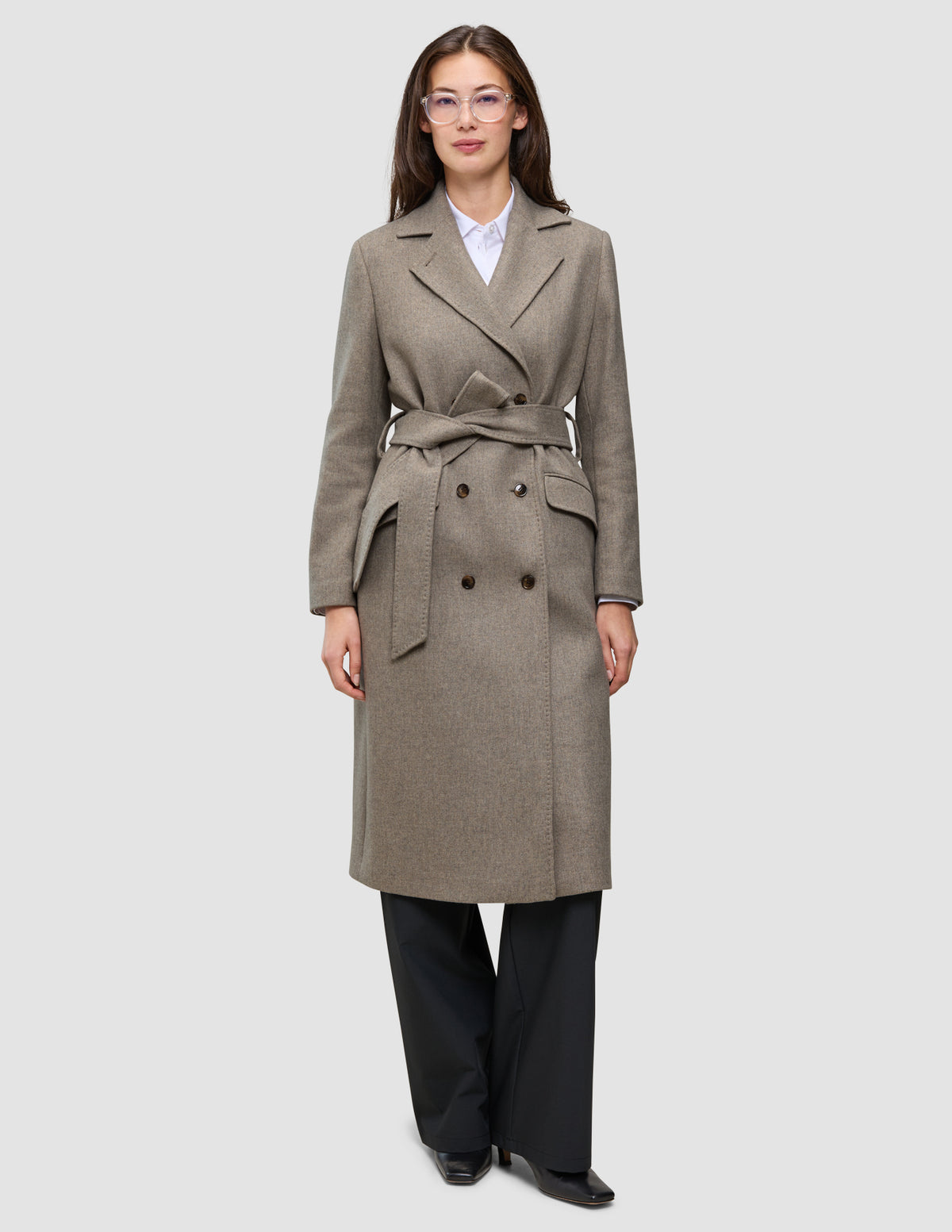 Eine Person, die einen graubeigen Damenmantel aus Wolle trägt – ein zweireihiges, mit einem Gürtel versehenes, minimalistisches skandinavisches Design –, dazu ein weißes Hemd mit Kragen und eine schwarze Hose.