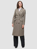 Eine Person, die einen Women Wool Coat Greige trägt – einen zweireihigen, knielangen, beigen Wollmantel mit einem passenden Bindegürtel –, dazu ein weißes Hemd und eine schwarze Hose, die das klare skandinavische Design unterstreichen.