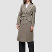 Eine Person, die einen Women Wool Coat Greige trägt – einen zweireihigen, knielangen, beigen Wollmantel mit einem passenden Bindegürtel –, dazu ein weißes Hemd und eine schwarze Hose, die das klare skandinavische Design unterstreichen.