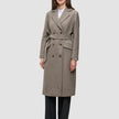 Eine Frau trägt einen Women Wool Coat Greige – einen zweireihigen Wollmantel mit Gürtel – über einem weißen Hemd und einer schwarzen Hose.