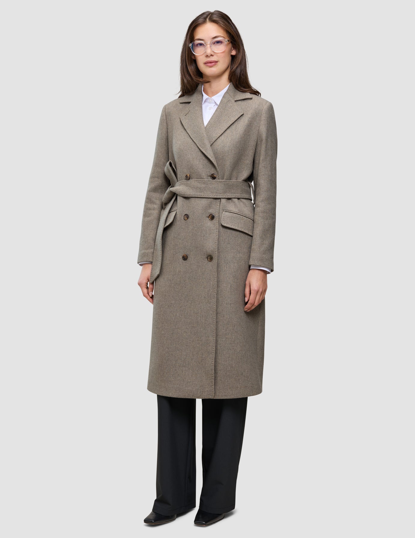 Eine Frau trägt einen Women Wool Coat Greige – einen zweireihigen Wollmantel mit Gürtel – über einem weißen Hemd und einer schwarzen Hose.