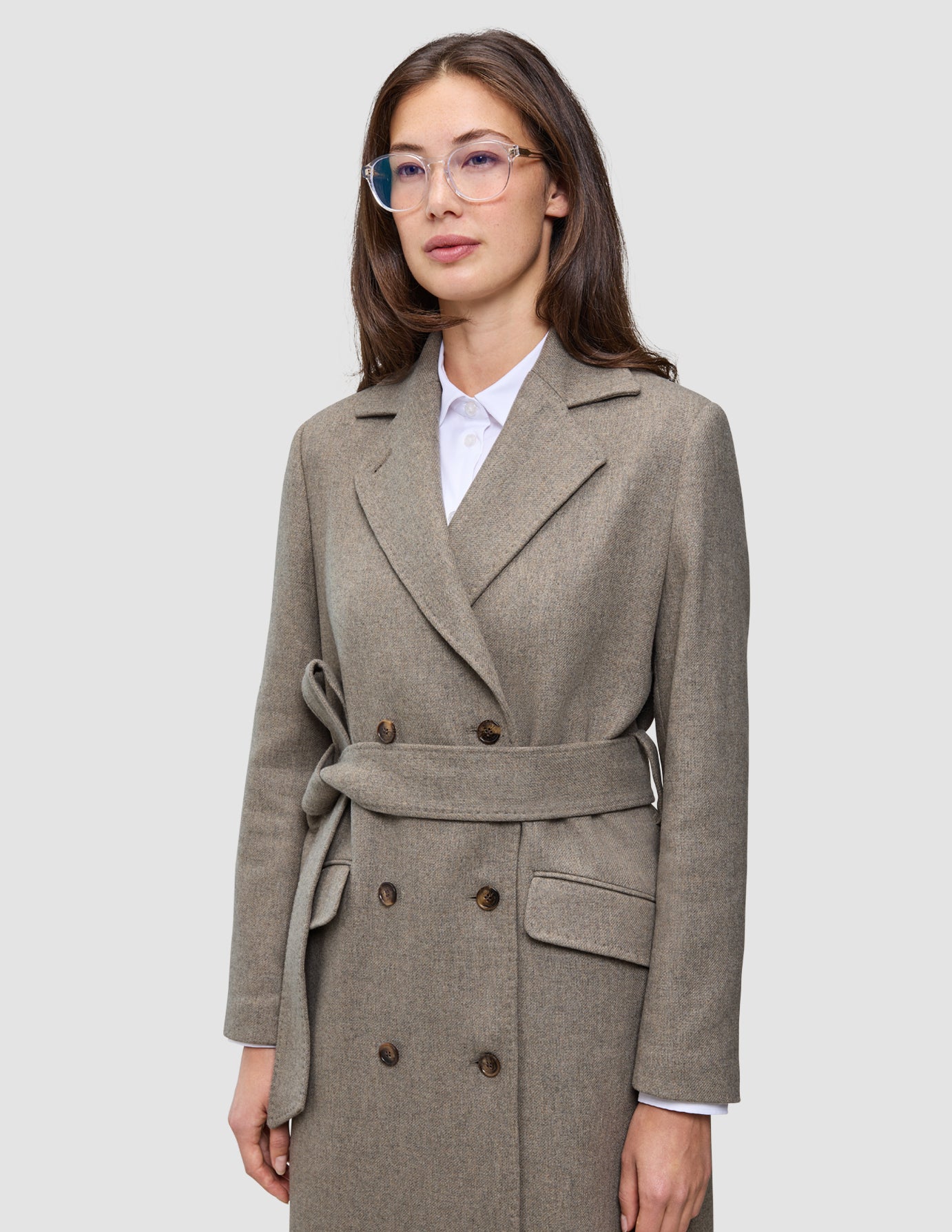 Eine Person, die einen Damen-Wollmantel in der Farbe Greige im doppelreihigen, mit einem Gürtel versehenen, minimalistischen skandinavischen Design über einem weißen Hemd mit Kragen trägt.