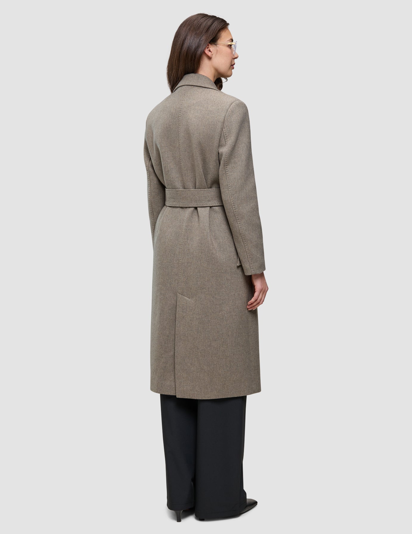Eine Person, von hinten gesehen, die einen Women Wool Coat Greige trägt – einen langen, mit einem Gürtel versehenen, greigefarbenen Wollmantel im skandinavischen Design – über einer schwarzen Hose.