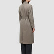 Eine Person, von hinten gesehen, die einen Women Wool Coat Greige trägt – einen langen, mit einem Gürtel versehenen, greigefarbenen Wollmantel im skandinavischen Design – über einer schwarzen Hose.
