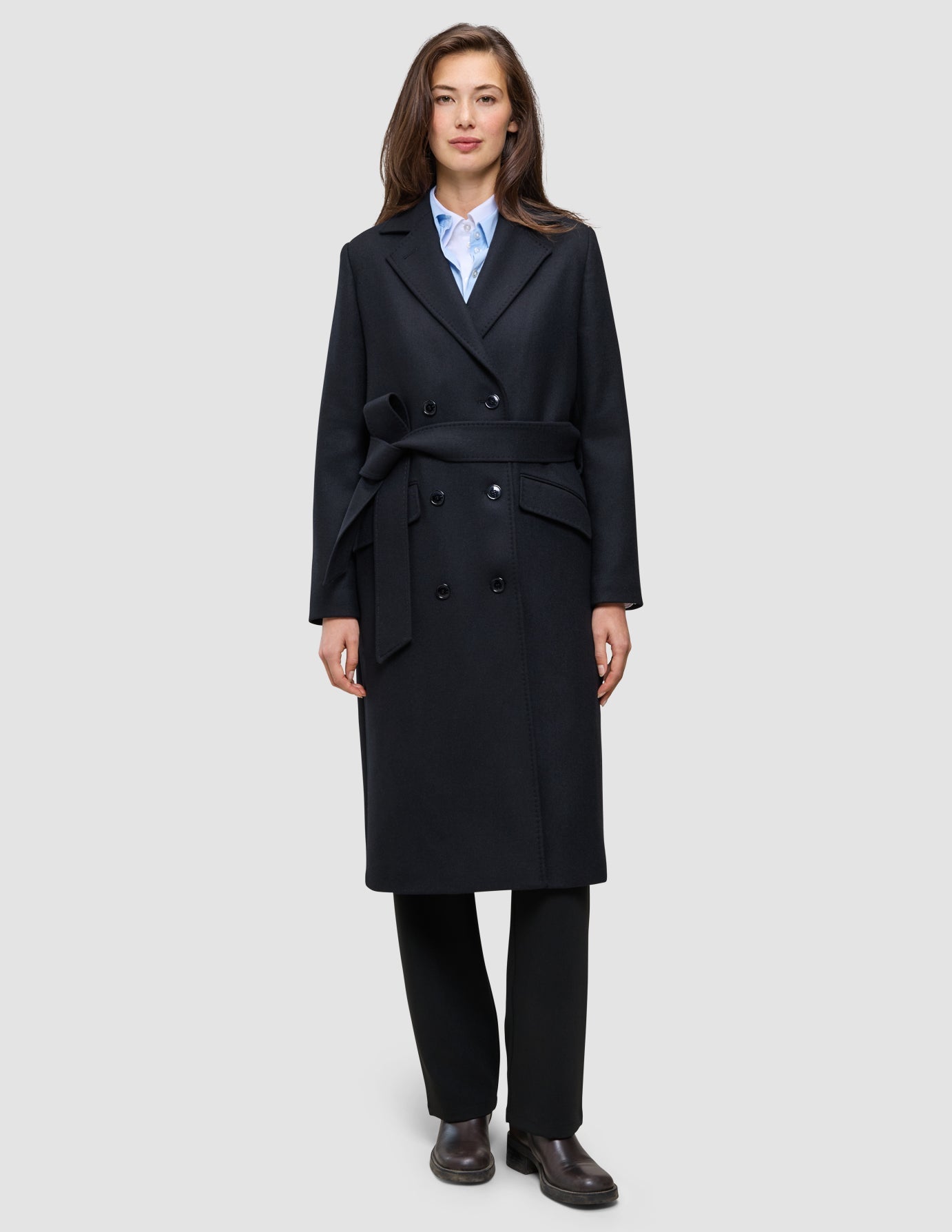 Eine Frau trägt einen schwarzen Damen-Wollmantel – einen langen, zweireihigen Wollmantel mit Gürtel in der Taille – über einem hellblauen Hemd mit Kragen und einer schwarzen Hose im klassischen skandinavischen Design.