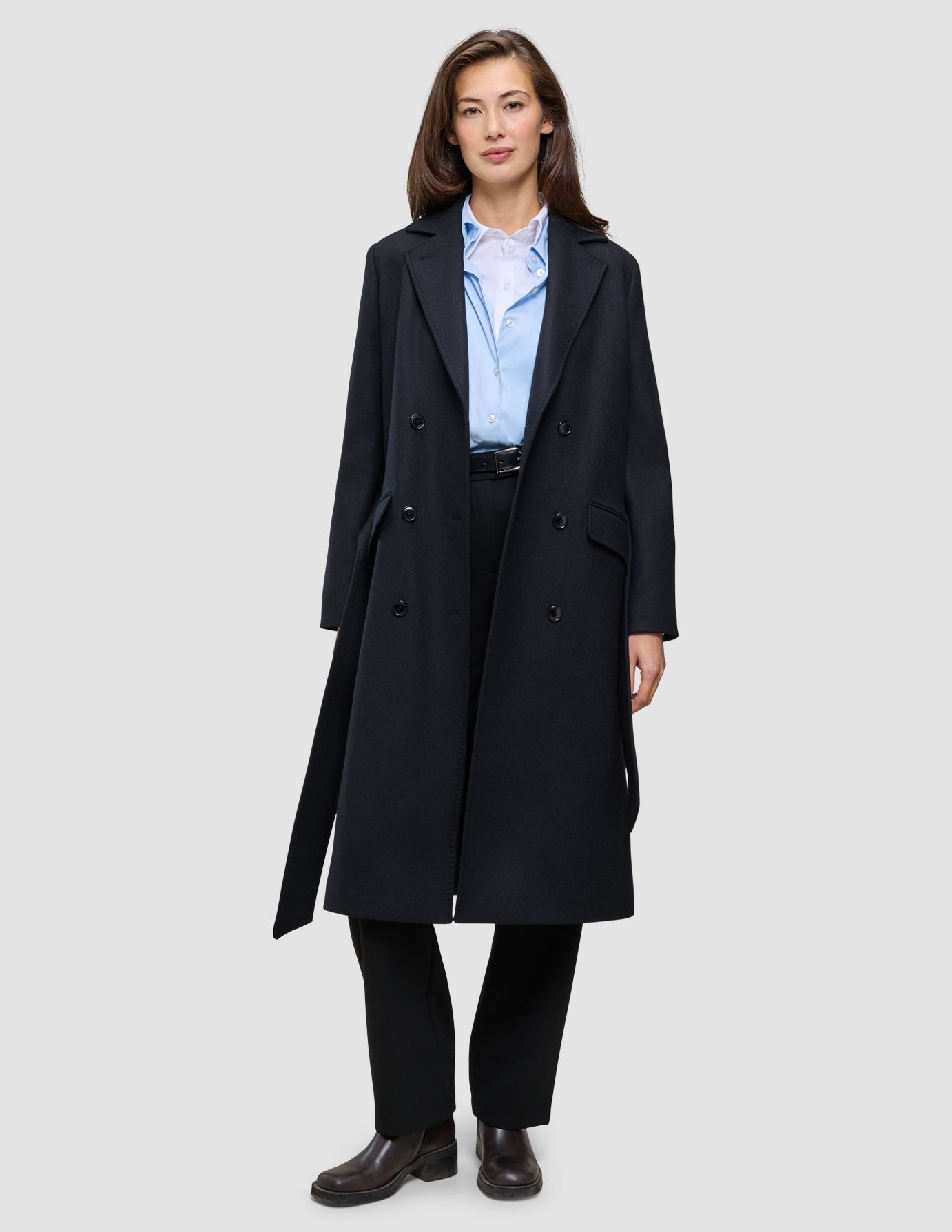 Eine Frau, die einen schwarzen Damenmantel aus Wolle über einem hellblauen Hemd mit Knöpfen und einer schwarzen Hose trägt, was das minimalistische skandinavische Design widerspiegelt.