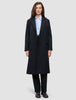 Eine Frau trägt einen schwarzen Damen-Wollmantel in knielanger Länge im Zweireiher-Stil über einem hellblauen Hemd mit Knöpfen und einer schwarzen Hose, was das klare skandinavische Design unterstreicht.