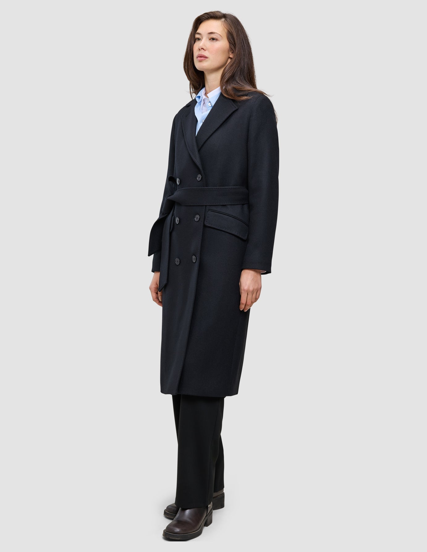 Eine Person, die einen schwarzen Damenmantel aus Wolle mit Gürtel an der Taille trägt, dazu ein hellblaues Hemd mit Kragen und eine schwarze Hose im schlichten, skandinavischen Design.