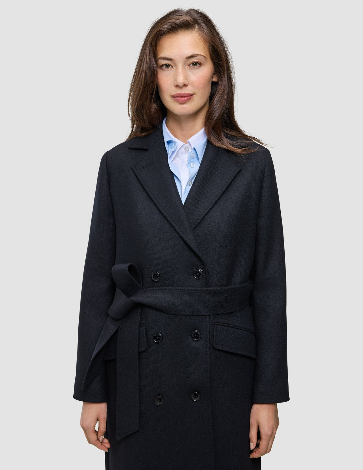 Eine Frau trägt einen schwarzen Damen-Wollmantel im Zweireiher-Stil mit Bindegürtel über einem hellblauen Hemd mit Kragen im skandinavischen Design.