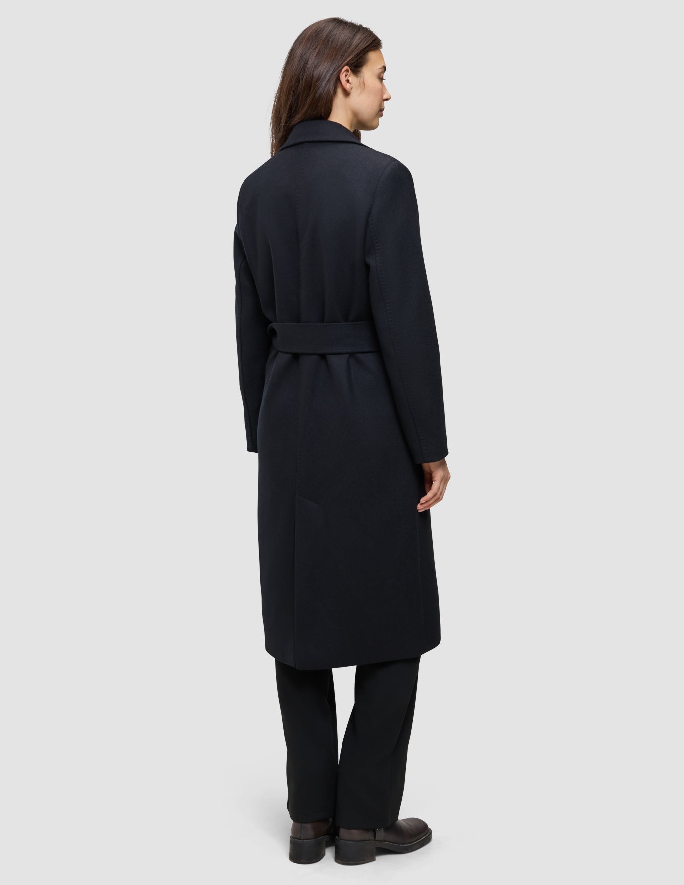 Eine Frau wird von hinten gezeigt. Sie trägt einen schwarzen Damenmantel aus Wolle in minimalistischem skandinavischem Design mit einer langen Silhouette und Gürtel.